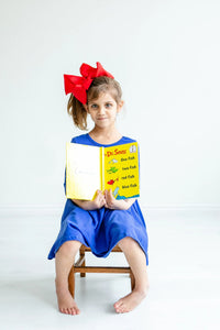 Royal blue twirl dress for girls, 3 4 sleeve pocket dress worn by young girl with red bow reading a Dr Seuss book