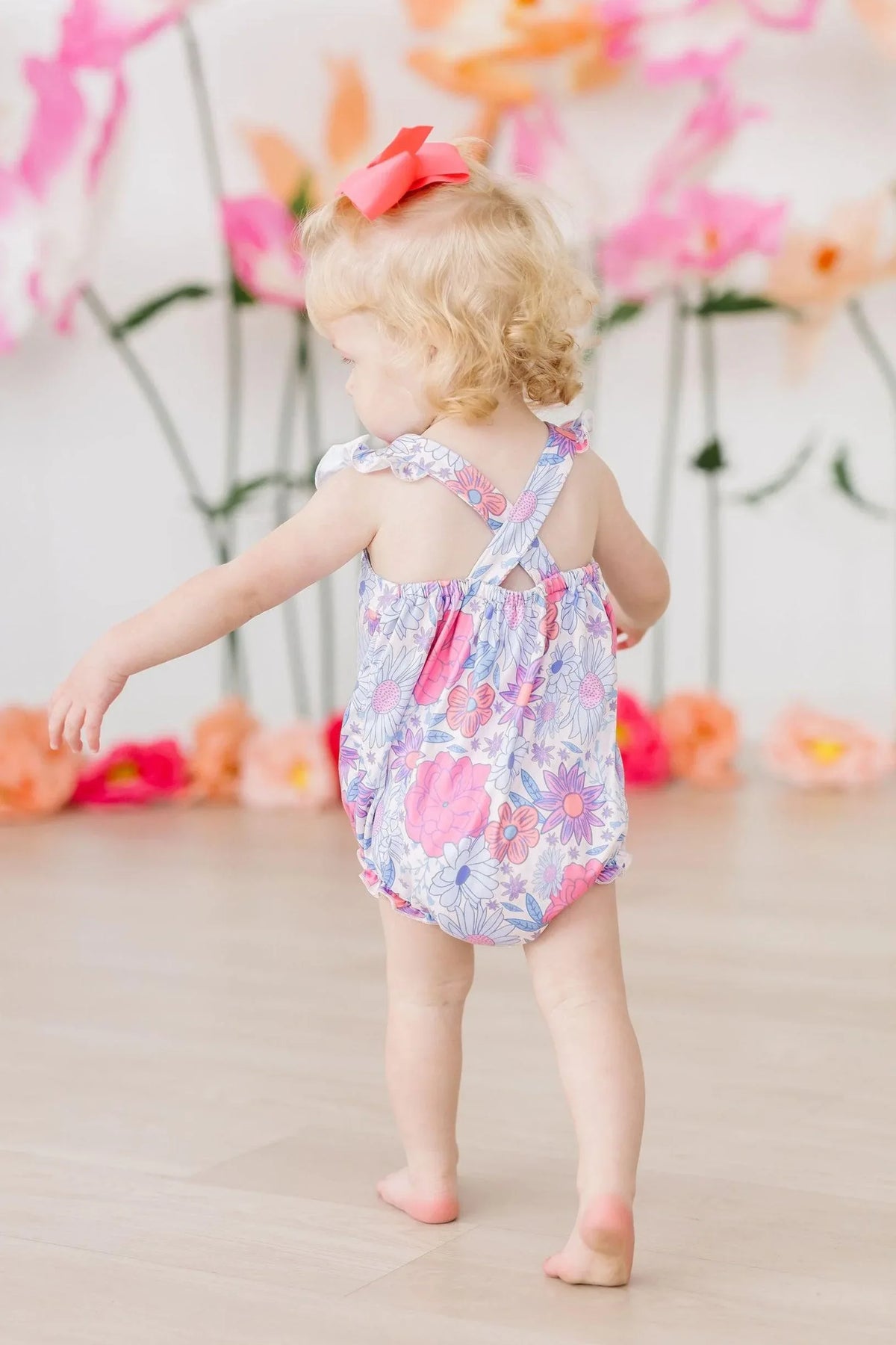 toddler girl wearing floral ruffle cross back romper walking indoors with pink flowers in background