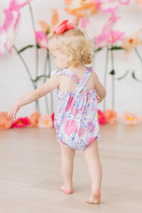 toddler girl wearing floral ruffle cross back romper walking indoors with pink flowers in background