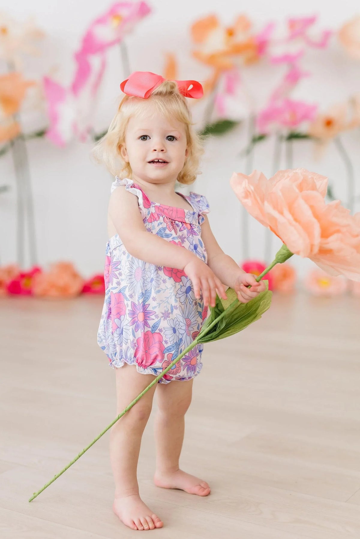 Toddler girl wearing floral ruffle cross back romper with pink bow holding large peach flower