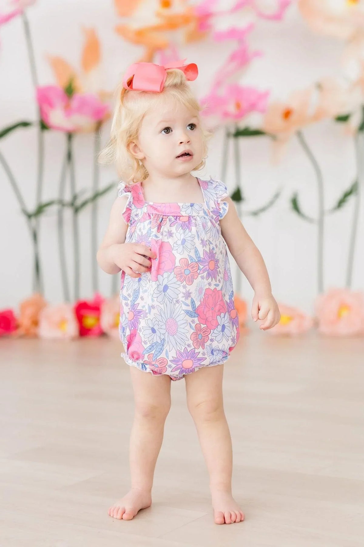 toddler girl wearing pink and purple floral ruffle cross back romper standing indoors with bow in hair