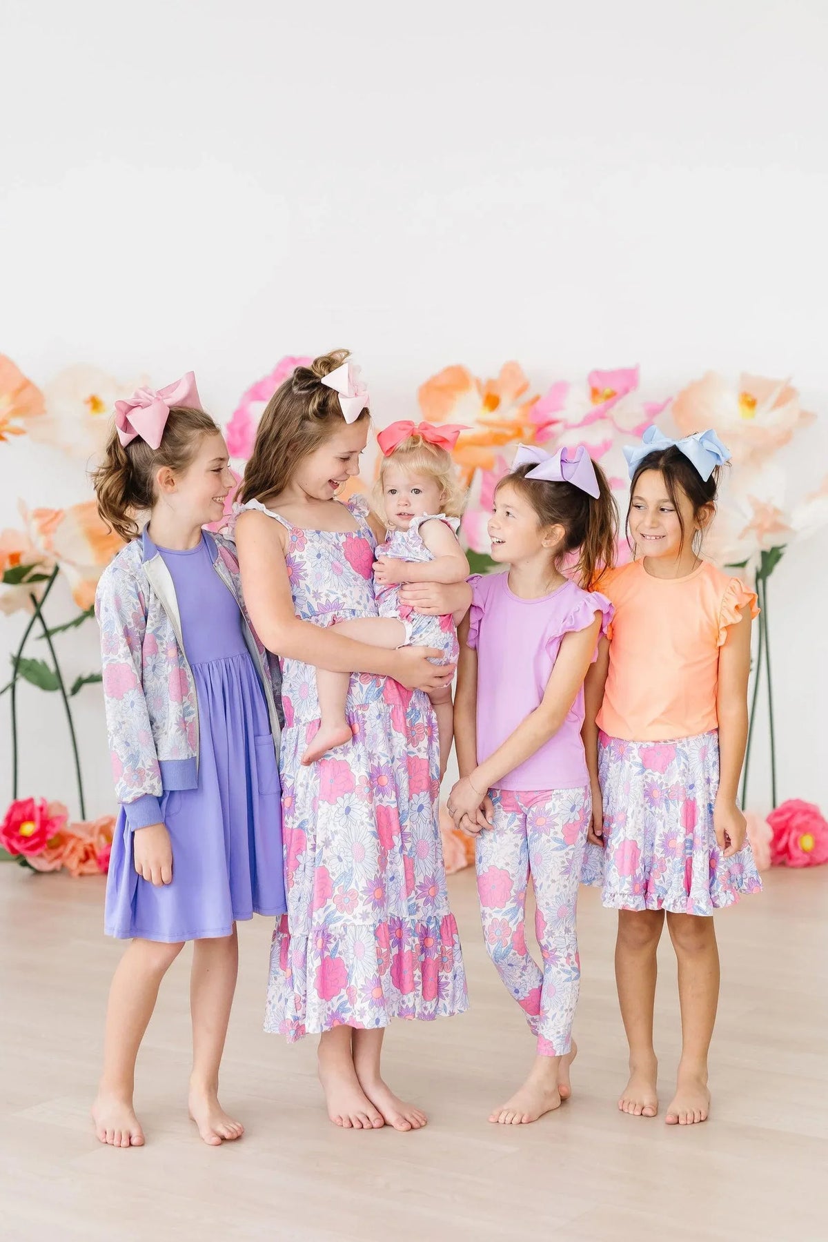 Four girls and one toddler wearing colorful floral and pastel outfits including a ruffle cross back romper standing together barefoot indoors