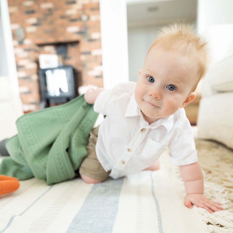 A happy baby playing on the floor with the Sage Knit Blanket by Goosewaddle®, highlighting its cozy design and gentle comfort.