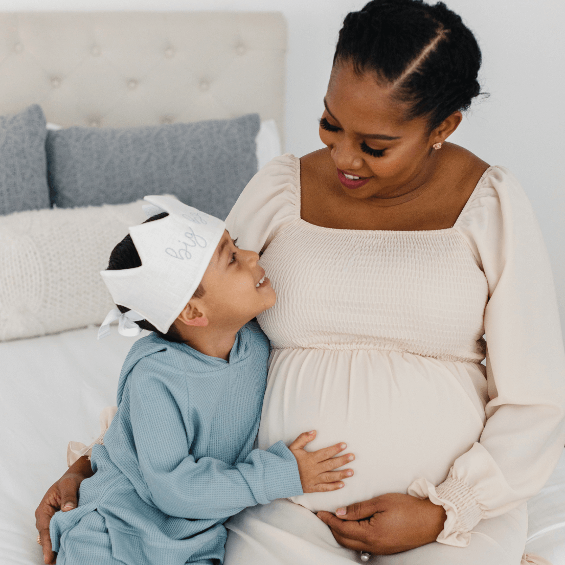Pregnant woman with a child wearing a baby bunting hat on a bed.