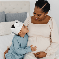Pregnant woman with a child wearing a baby bunting hat on a bed.