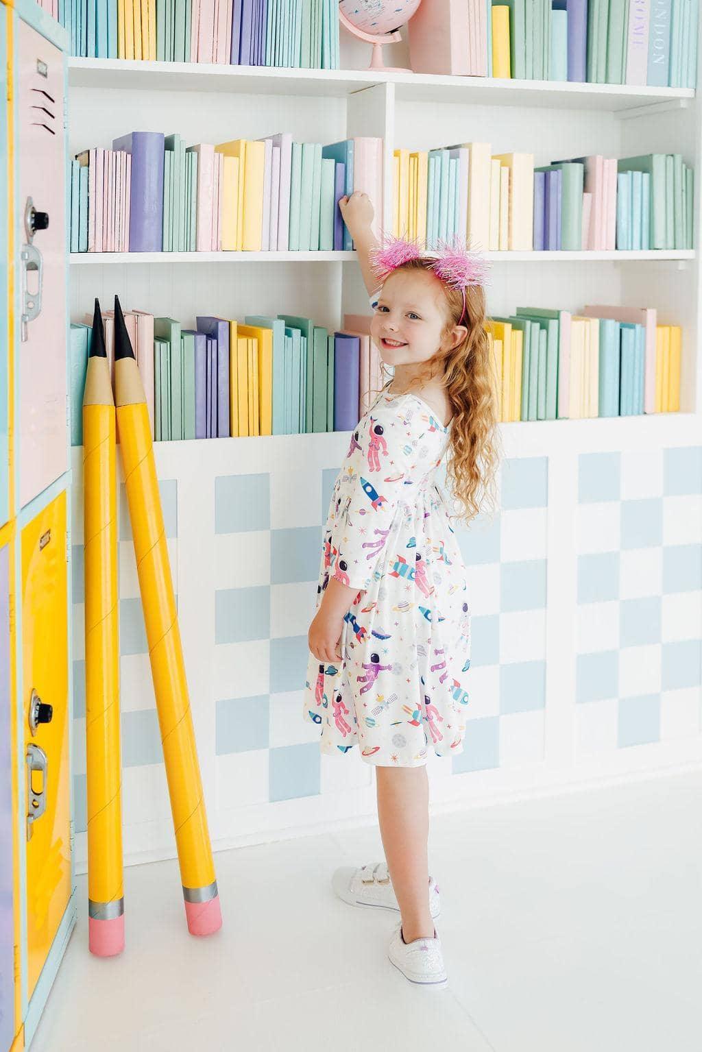 girl wearing a white space print twirl dress for girls reaching for a book in a colorful library