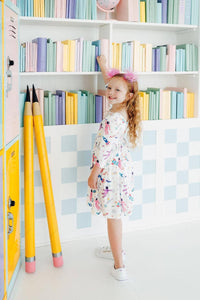 girl wearing a white space print twirl dress for girls reaching for a book in a colorful library