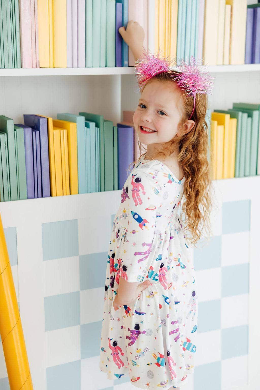 young girl wearing white space print twirl dress for girls reaching for books on shelf