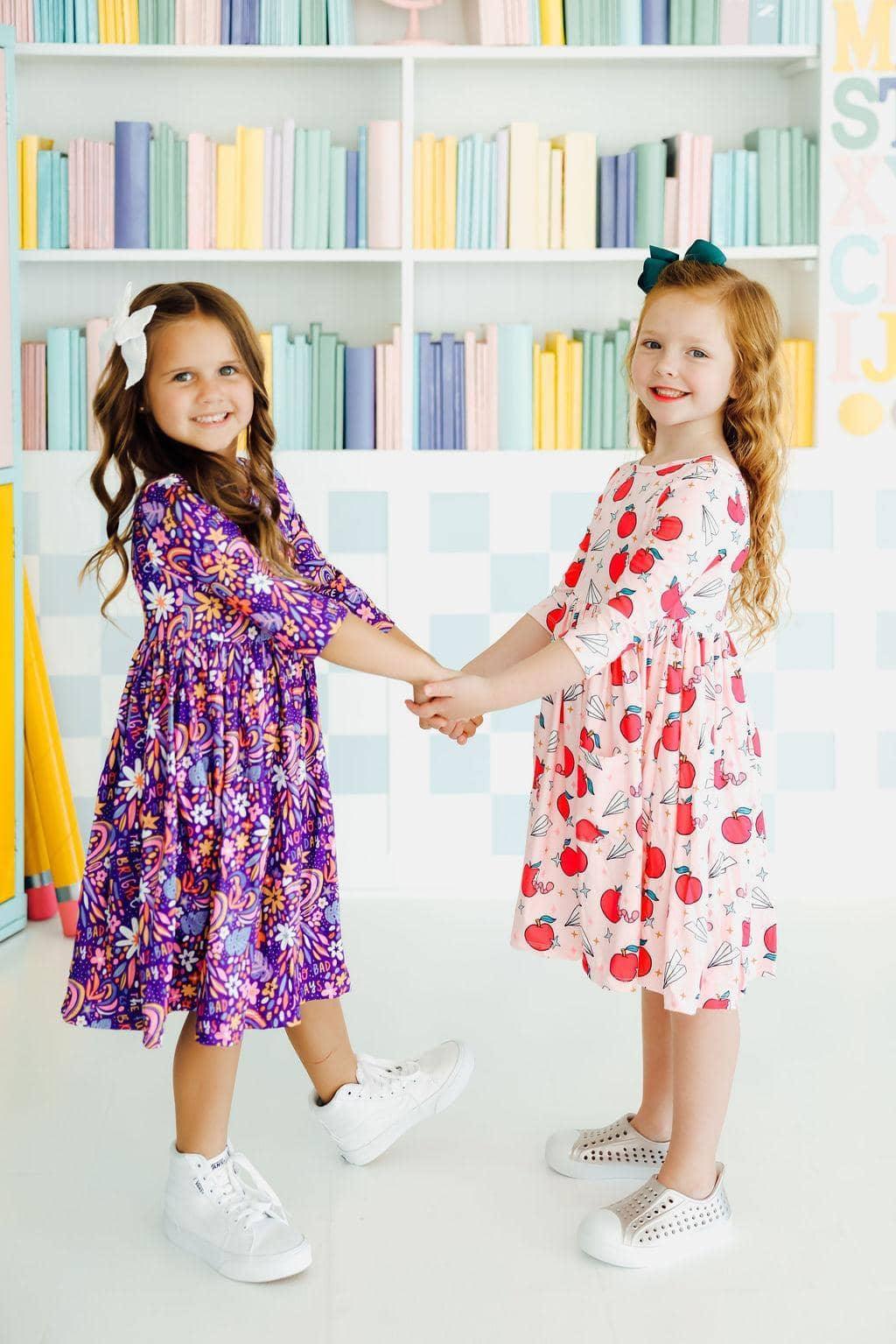 Two girls wearing Mila & Rose star student twirl dress for girls, one in purple floral and one in white with red apples, 3 4 sleeve