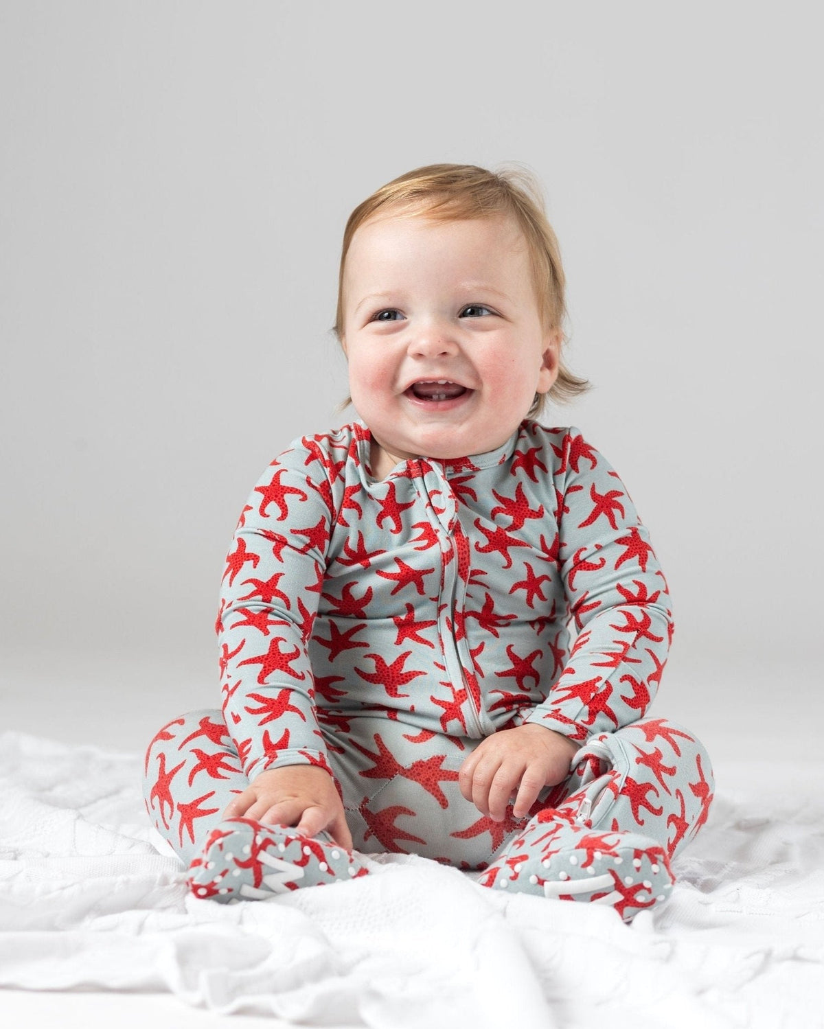 Happy baby wearing gray bamboo zipper footie pajamas with red starfish print and gripped soles.