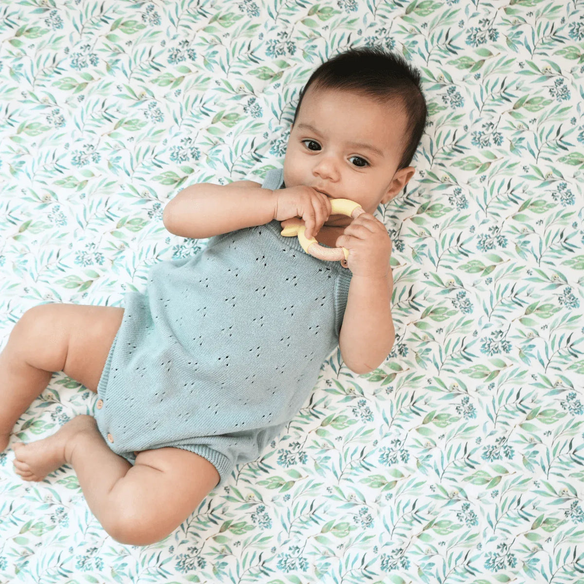 Baby lying on white and green floral super soft baby play mat wearing light blue knitted romper chewing on teething ring