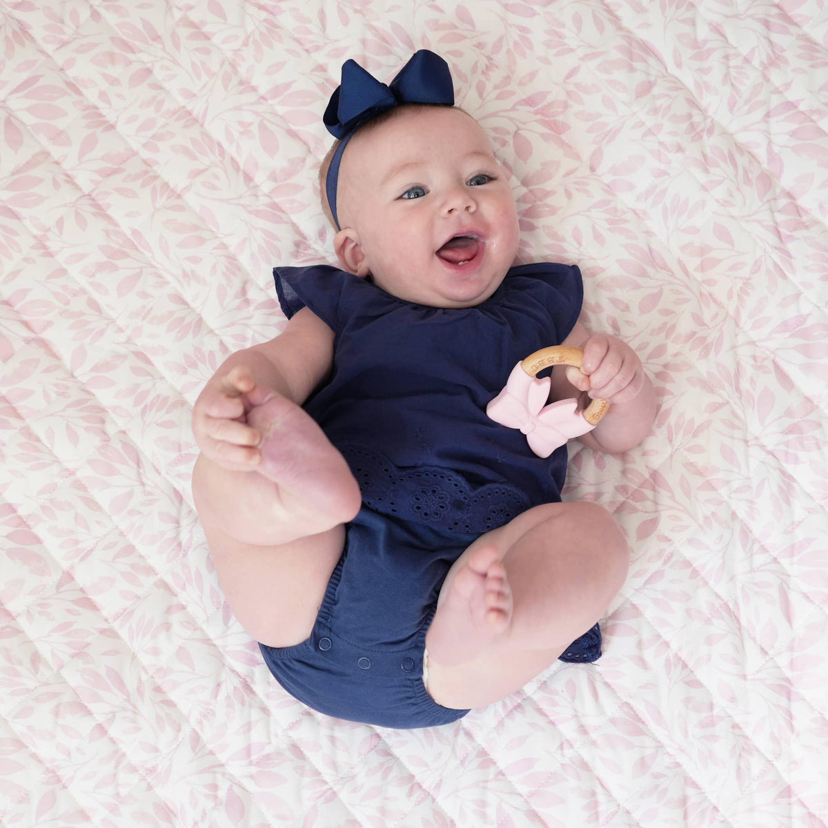 smiling baby girl wearing navy blue outfit and headband lying on pink super soft baby play mat holding teether