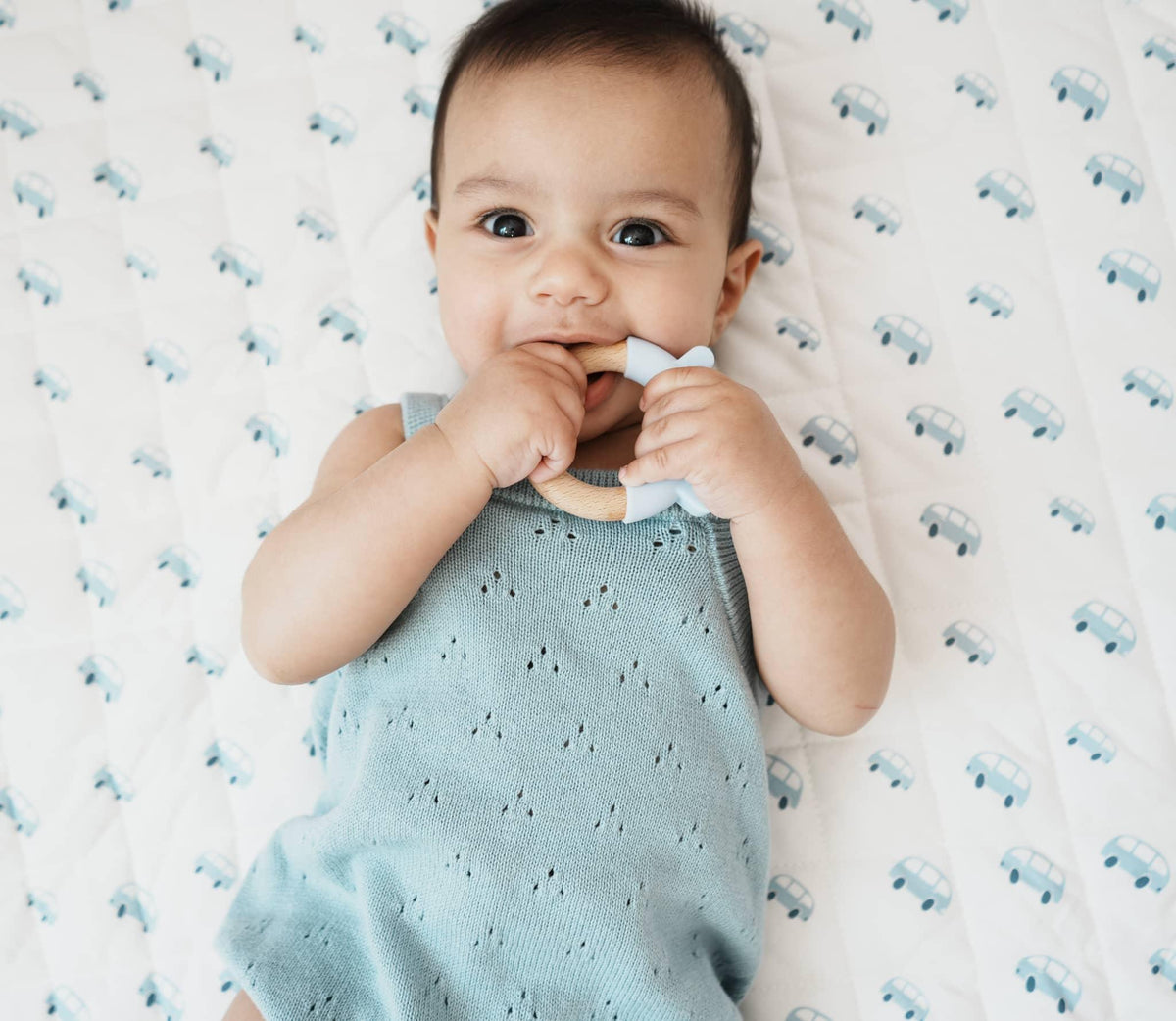 Baby lying on white floor mat with blue car prints holding teething toy on super soft baby play mat
