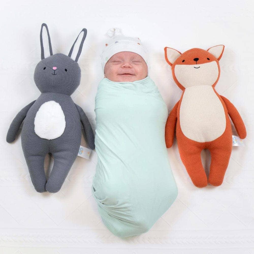 Newborn baby swaddled in light green swaddle blanket next to gray bunny and orange fox plush toys