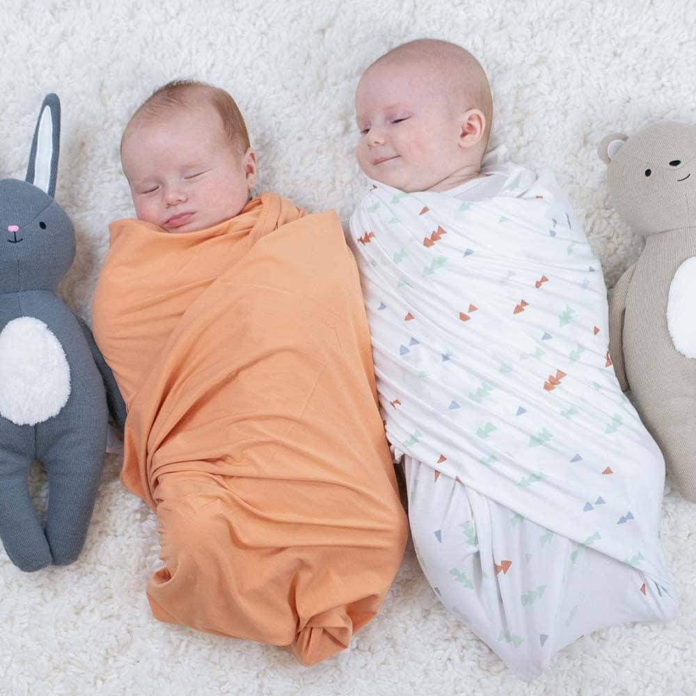 Two newborn babies wrapped in soft swaddle blankets one in solid orange and the other in white with colorful prints