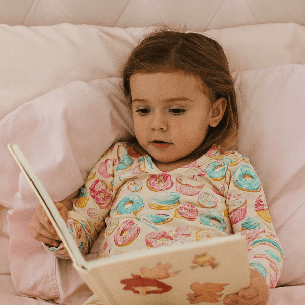 Child in pajamas reading a book on a pink bed