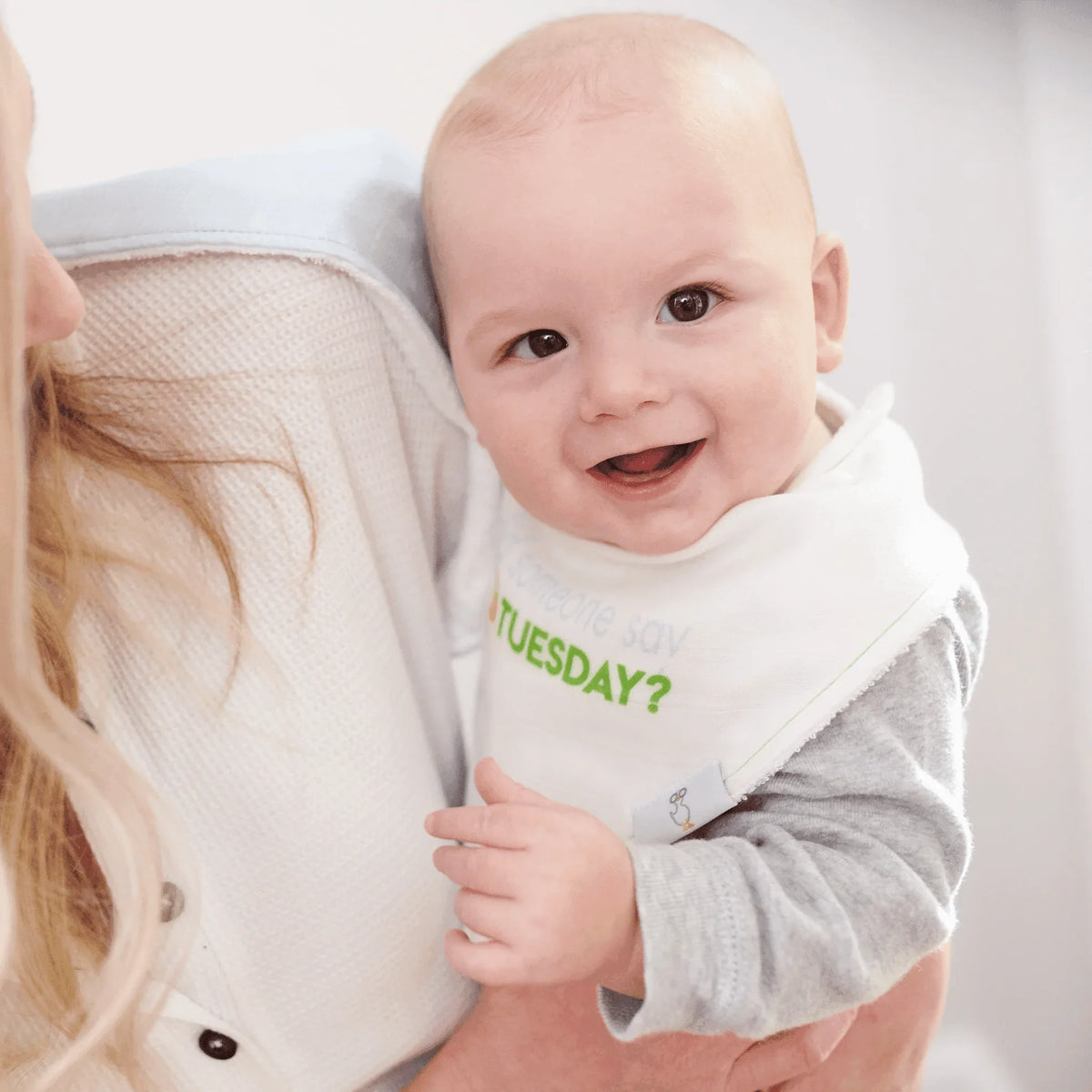 smiling baby wearing white and green taco baby bib set held by adult in gray and white clothing