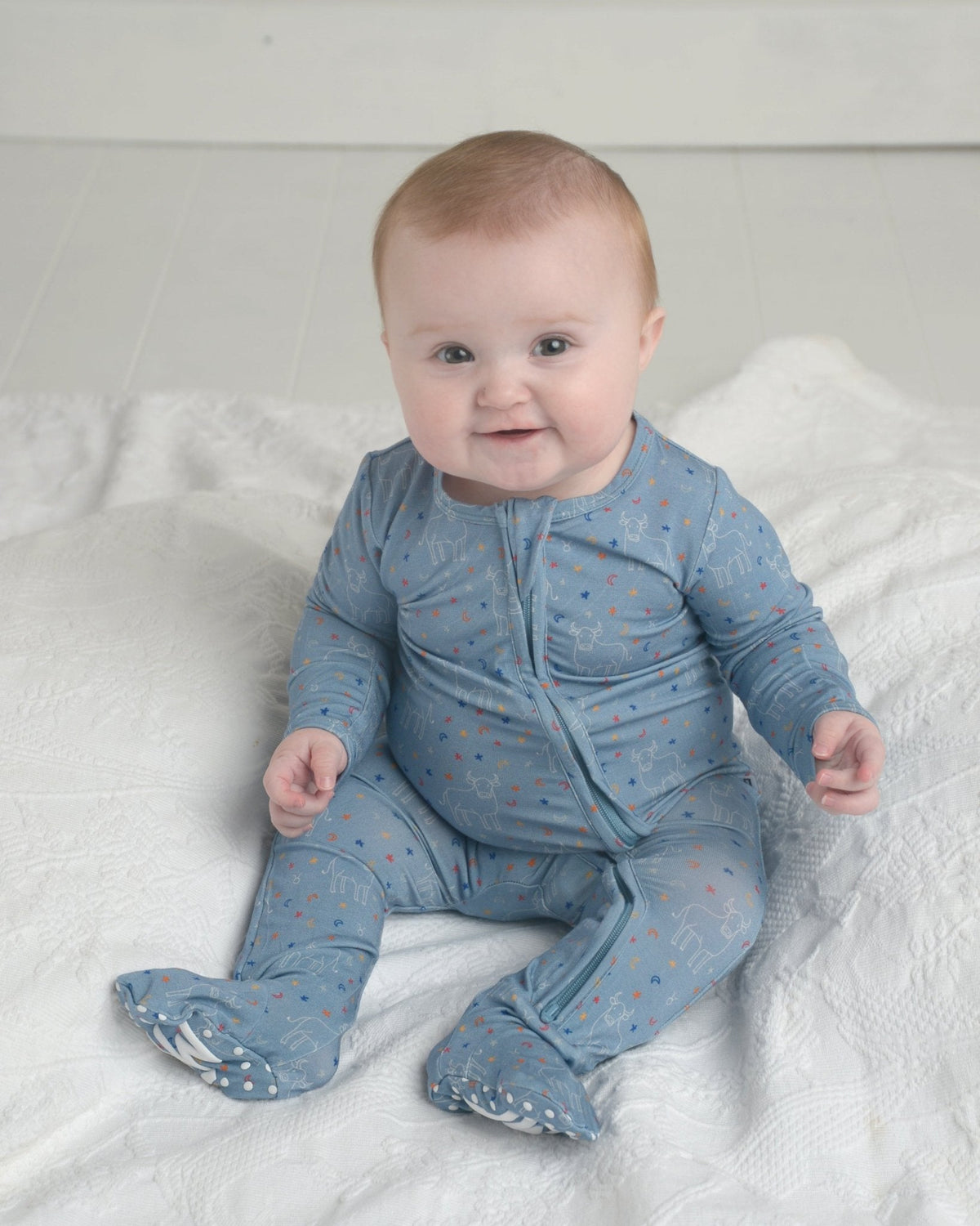 Baby wearing blue Taurus zodiac bamboo zipper footie pajamas with bull and star print, sitting on white blanket.