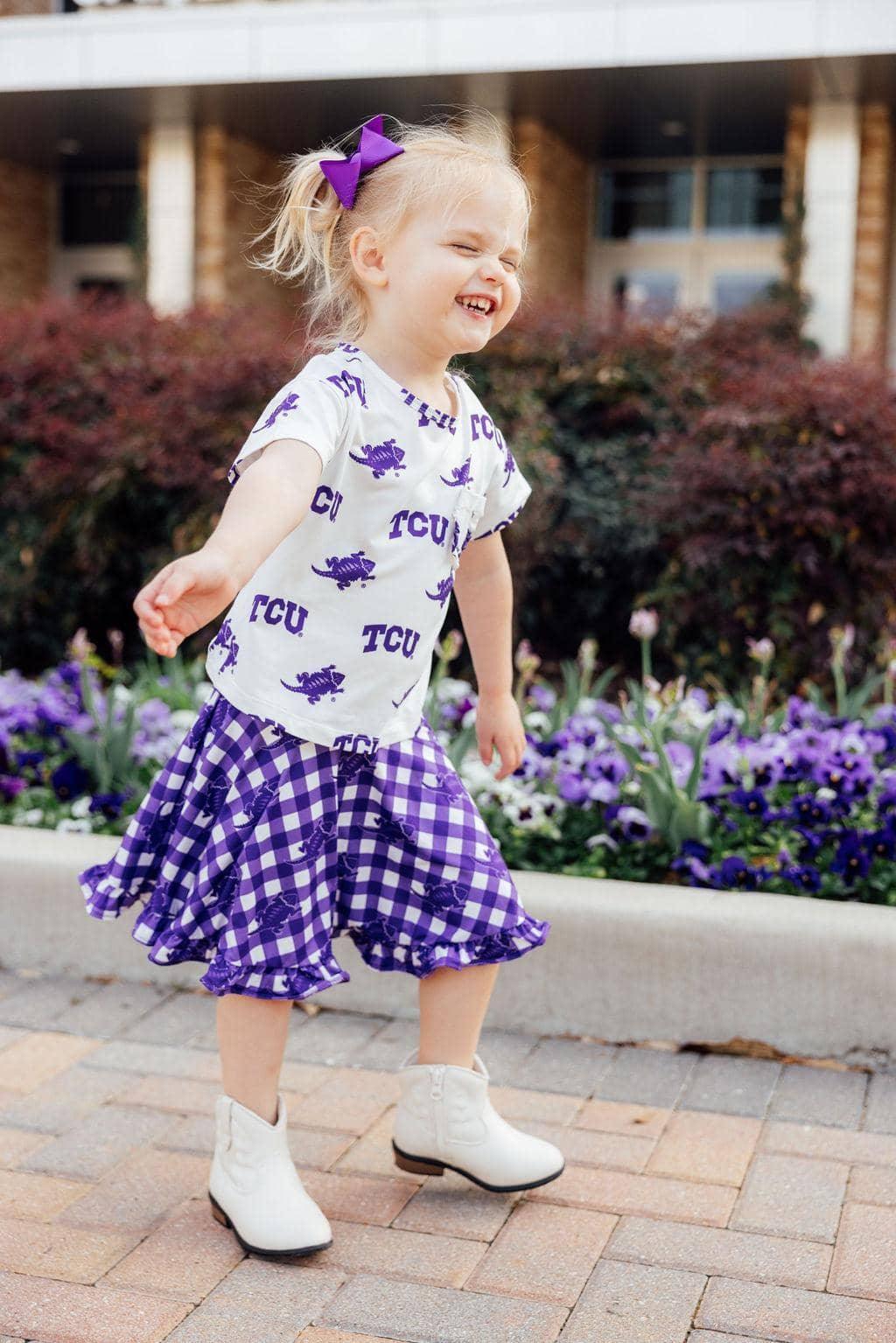 Toddler girl wearing purple checkered ruffle twirl skort and TCU print shirt with bow and white boots