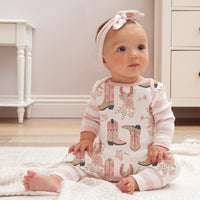 Baby girl in cream western romper with pink cowboy boot print and striped sleeves sitting on white rug