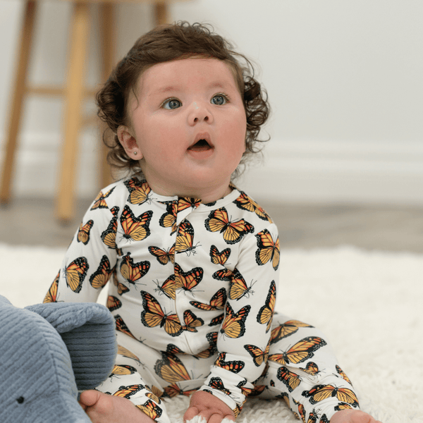 Baby wearing a onesie with butterfly pattern sitting on a carpeted floor.