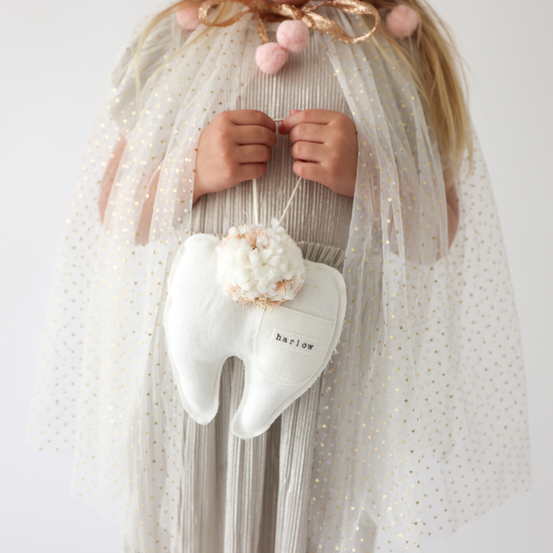 Child holding a white heart-shaped plush toy with 'harlow' on it, wearing a sheer white dress.