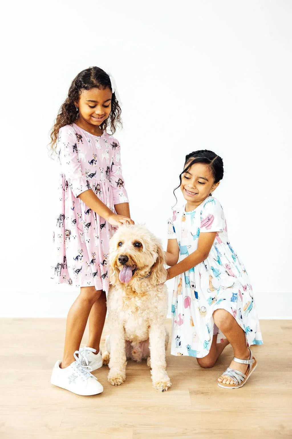 Two girls wearing pink and white twirl dress for girls playing with a fluffy dog indoors