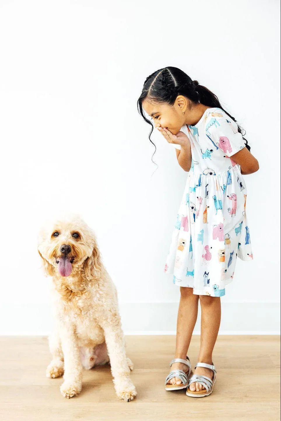 Young girl wearing white twirl dress for girls with colorful animal prints laughing and bending over next to a happy golden doodle dog