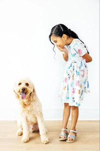 Young girl wearing white twirl dress for girls with colorful animal prints laughing and bending over next to a happy golden doodle dog