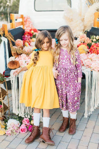 Two young girls wearing mustard yellow and purple floral twirl dress for girls standing and holding hands outdoors