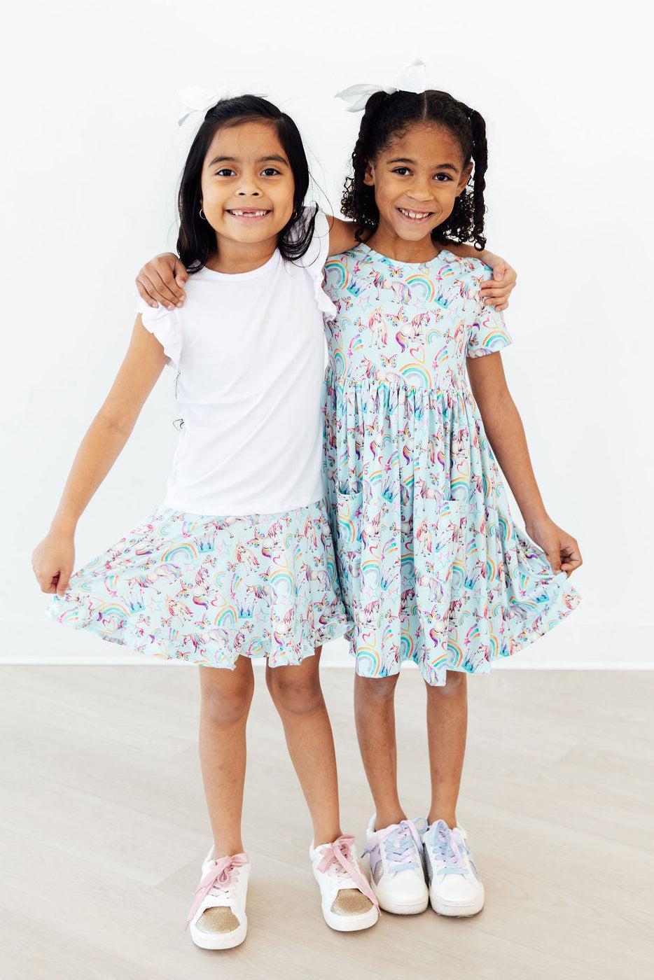 Two young girls smiling with arms around each other wearing unicorn dress for toddlers in pastel colors, twirling and posing