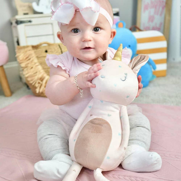 baby girl holding pink unicorn plush toy for babies sitting on pink blanket with white bow headband
