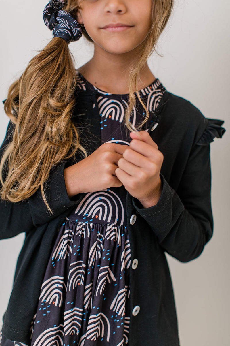 A girl in a Black Ruffle Cardigan, holding her hands together, showcasing the soft fabric and ruffle details, worn over a colorful dress, ideal for stylish comfort.