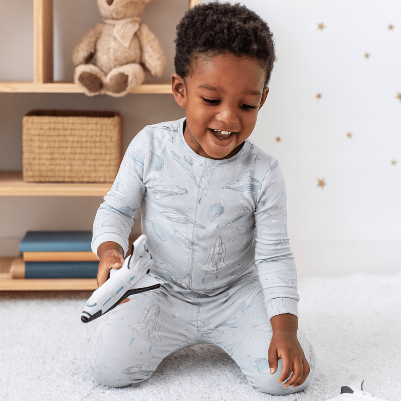 Happy kid in Up and Away Big Kid Pajama on a white rug, playing with a toy rocket.