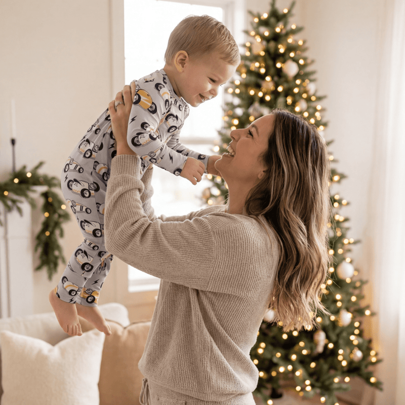 Baby wearing Vintage Racecars Convertible Romper lifted by mom under a Christmas tree in a cozy living room.
