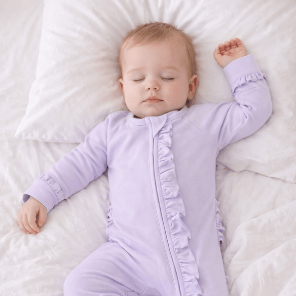 Baby wearing lavender Violets Are Violet Ruffle Footie with 2-way zipper and ruffles, sleeping on white sheets.