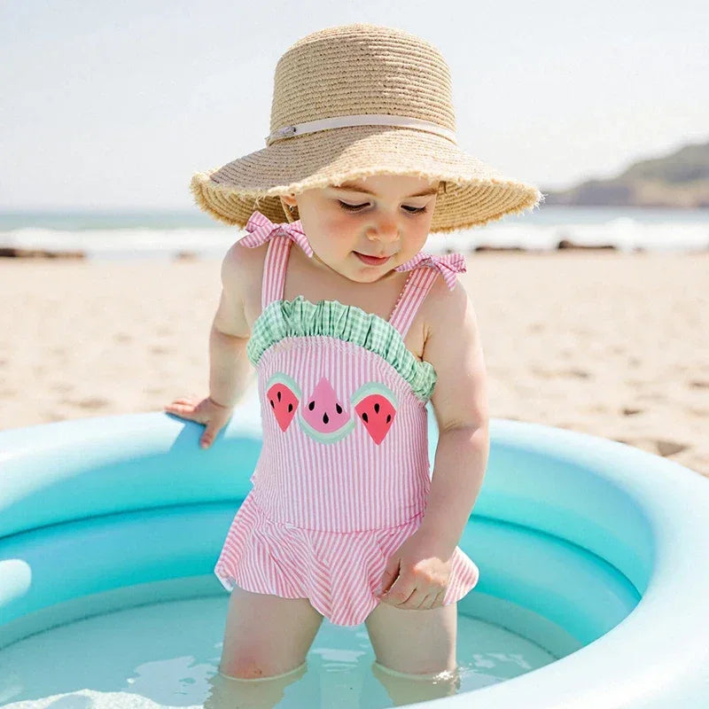 Pink and green cotton watermelon baby swimsuit with ruffled straps worn by toddler at the beach