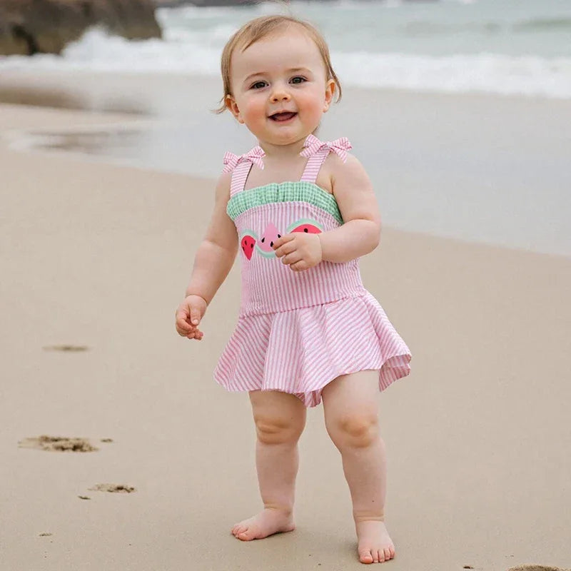 Pink and green cotton watermelon baby swimsuit with ruffled skirt worn by baby on the beach