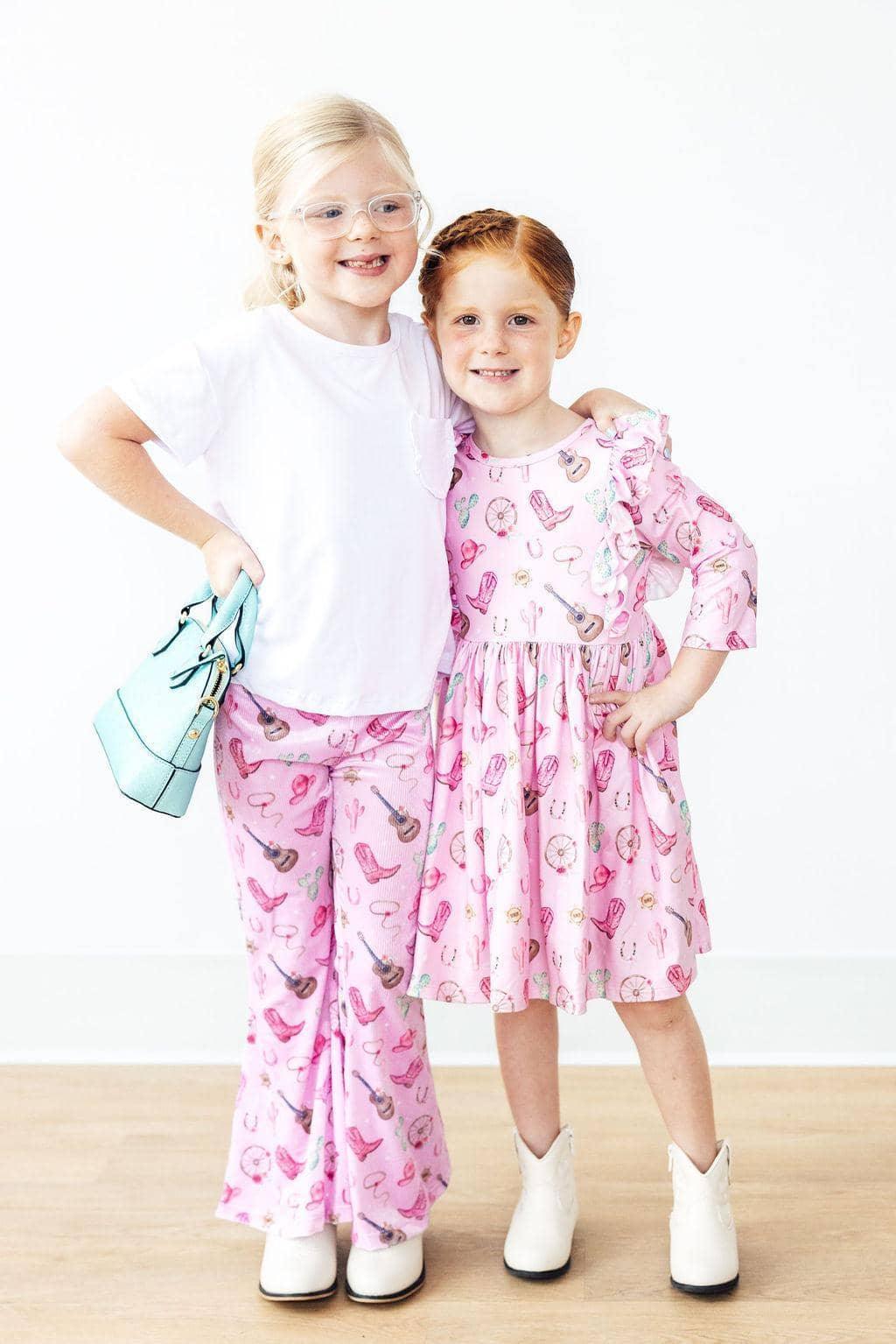 Two young girls standing, one wearing pink western dress for girls with ruffle sleeves, and the other in matching pink pants
