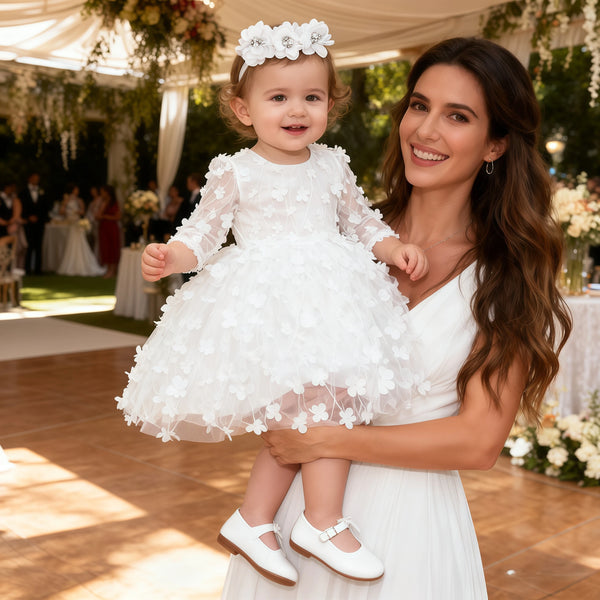 toddler girl wearing white flower girl dress with headband held by smiling woman at outdoor event