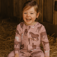 A young child wearing a pink long-sleeve zip-up sleeper with an allover horse and donkey print, sitting on a bed of straw inside a barn.