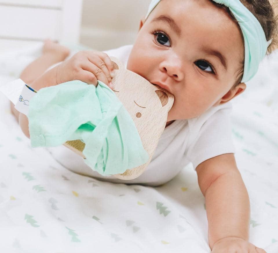 Baby girl wearing mint headband chewing on wooden baby teether and blanket set while lying on patterned blanket