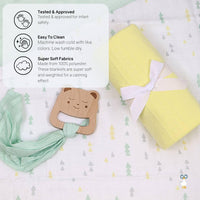 Rolled yellow knit baby blanket with white ribbon and wooden bear teether on a patterned baby sheet