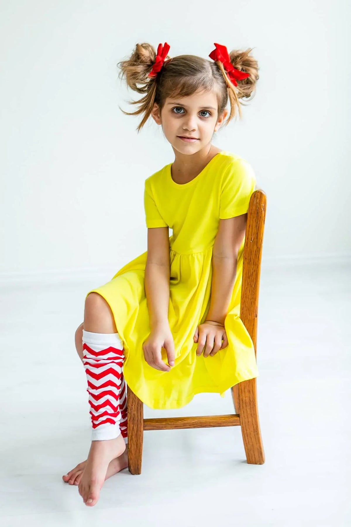 Young girl wearing yellow twirl dress for girls sitting on wooden chair with red hair bows