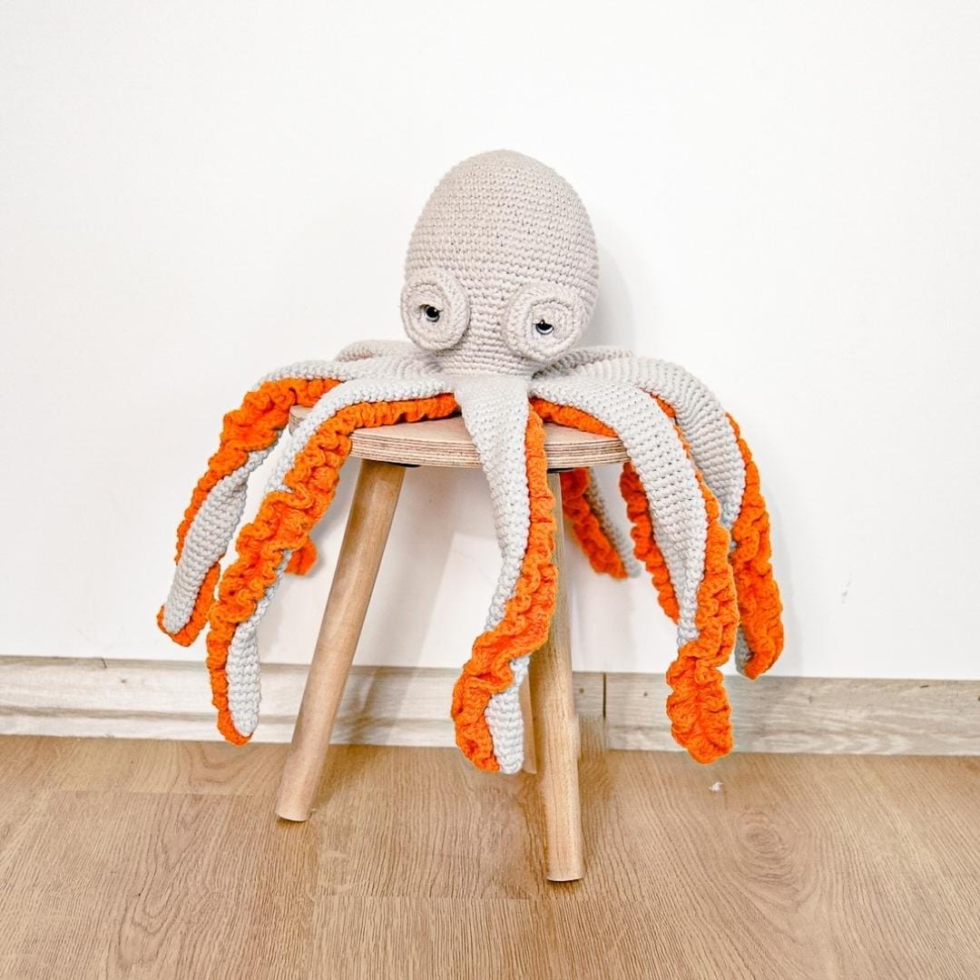 Grey crocheted octopus with bright orange curling arms sits on a small wooden stool. Perfect plush for nurseries.
