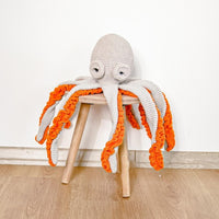 Grey crocheted octopus with bright orange curling arms sits on a small wooden stool. Perfect plush for nurseries.