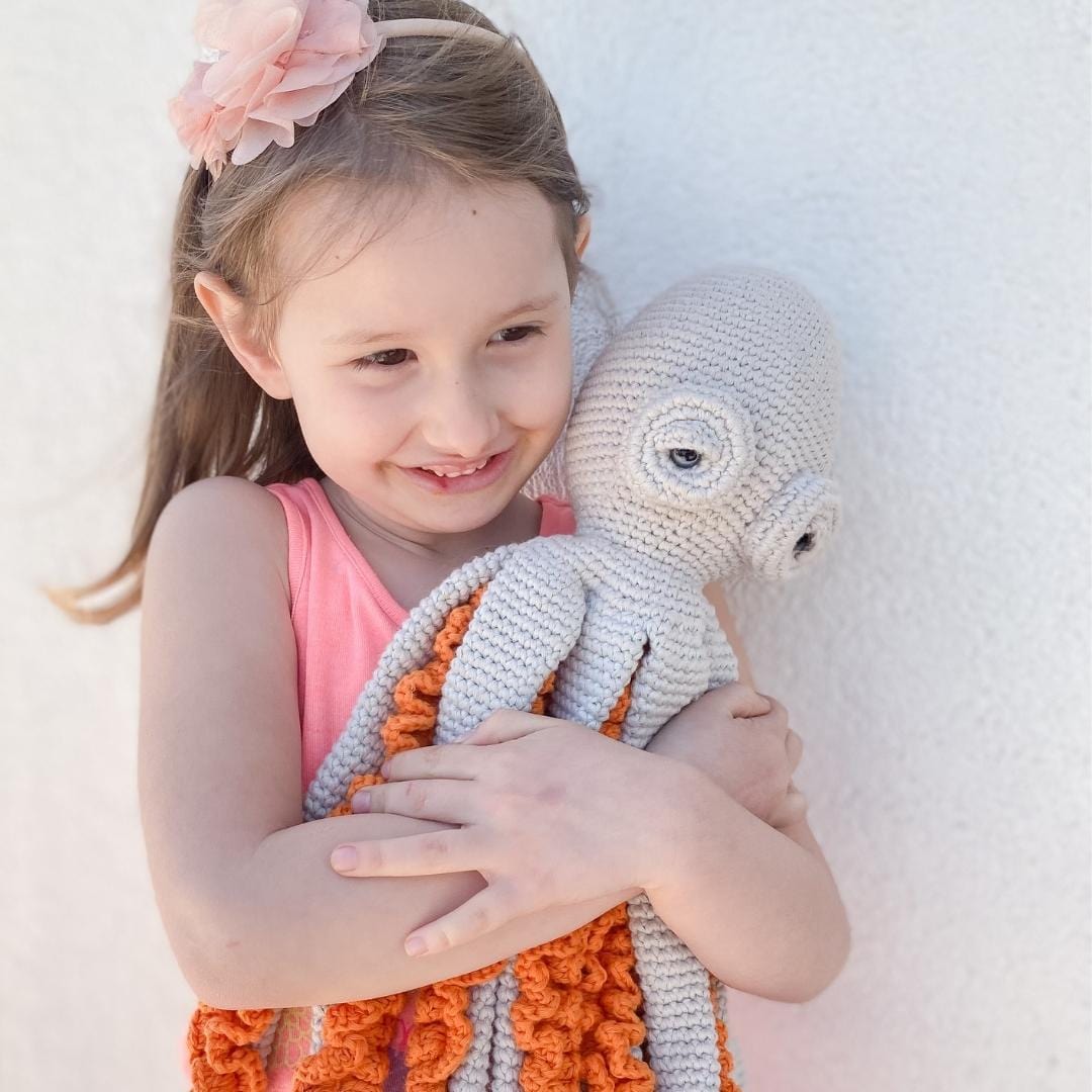Smiling girl holding grey crochet octopus with curling orange arms. Soft organic cotton toy for nursery or kids room adventure.