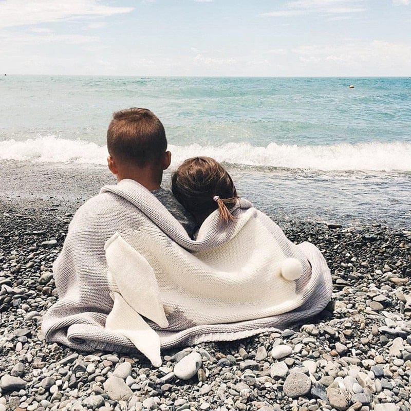Gray and white bunny baby blanket swaddle for infants with ear and tail details sitting on pebbled beach